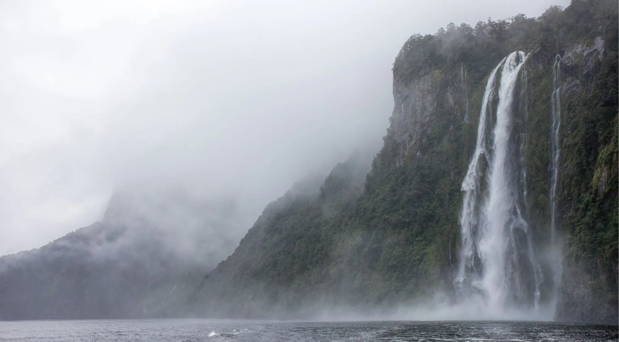 Waterfall pouring down a cliff face into Milford Sound with low cloud across the fiord