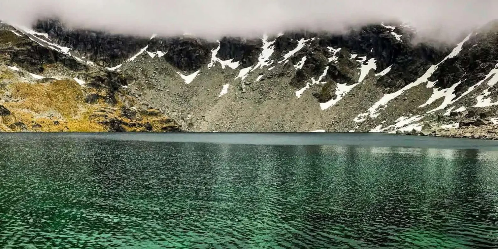 Green alpine tarn ringed by snow-streaked rock, seen from the air