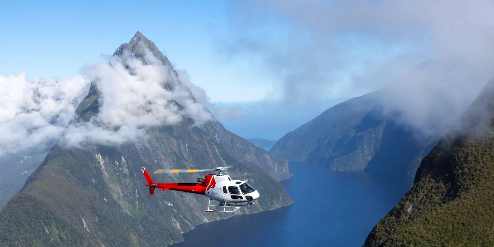 Helicopter flying past Mitre Peak with cloud spilling over the fiord walls
