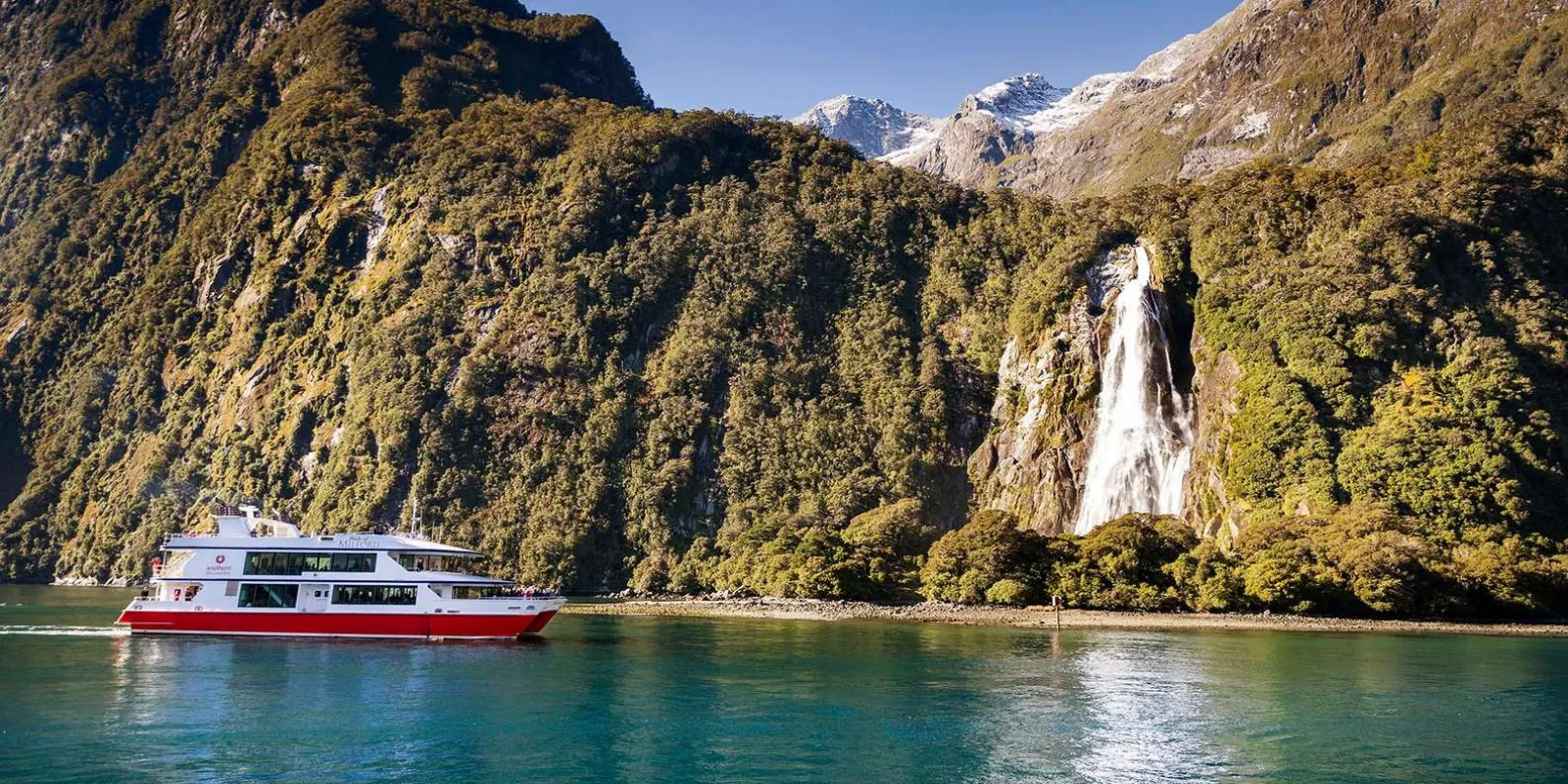 Cruise boat dwarfed by a waterfall pouring down the cliffs of Milford Sound